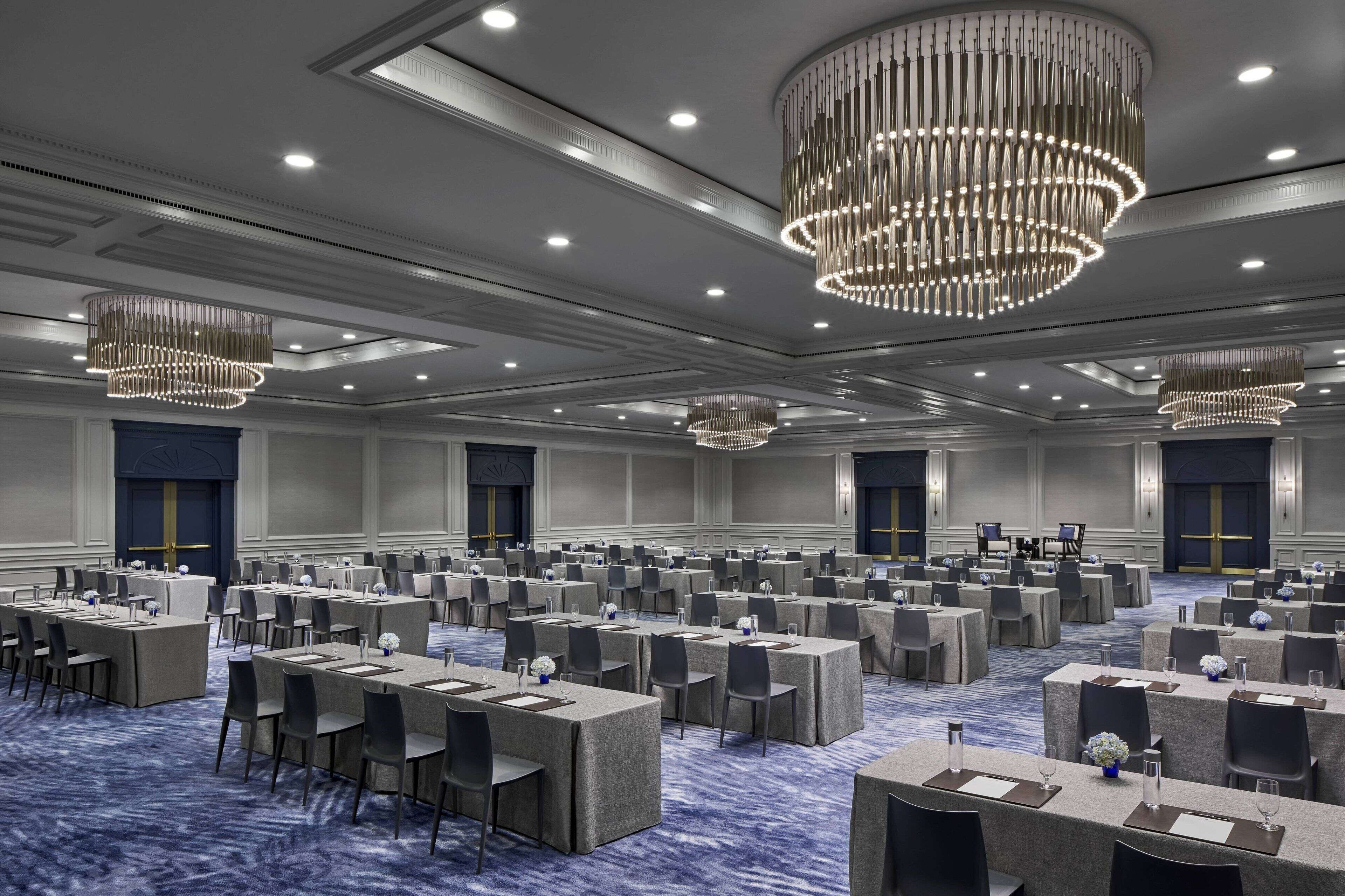 Une grande salle de conférence dotée de nombreuses tables et chaises, de lustres élégants et d’un sol recouvert d’une moquette bleue.
