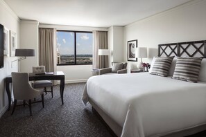 A hotel room with a large bed, a desk, a chair, a lamp, and a view of the city through a large window.