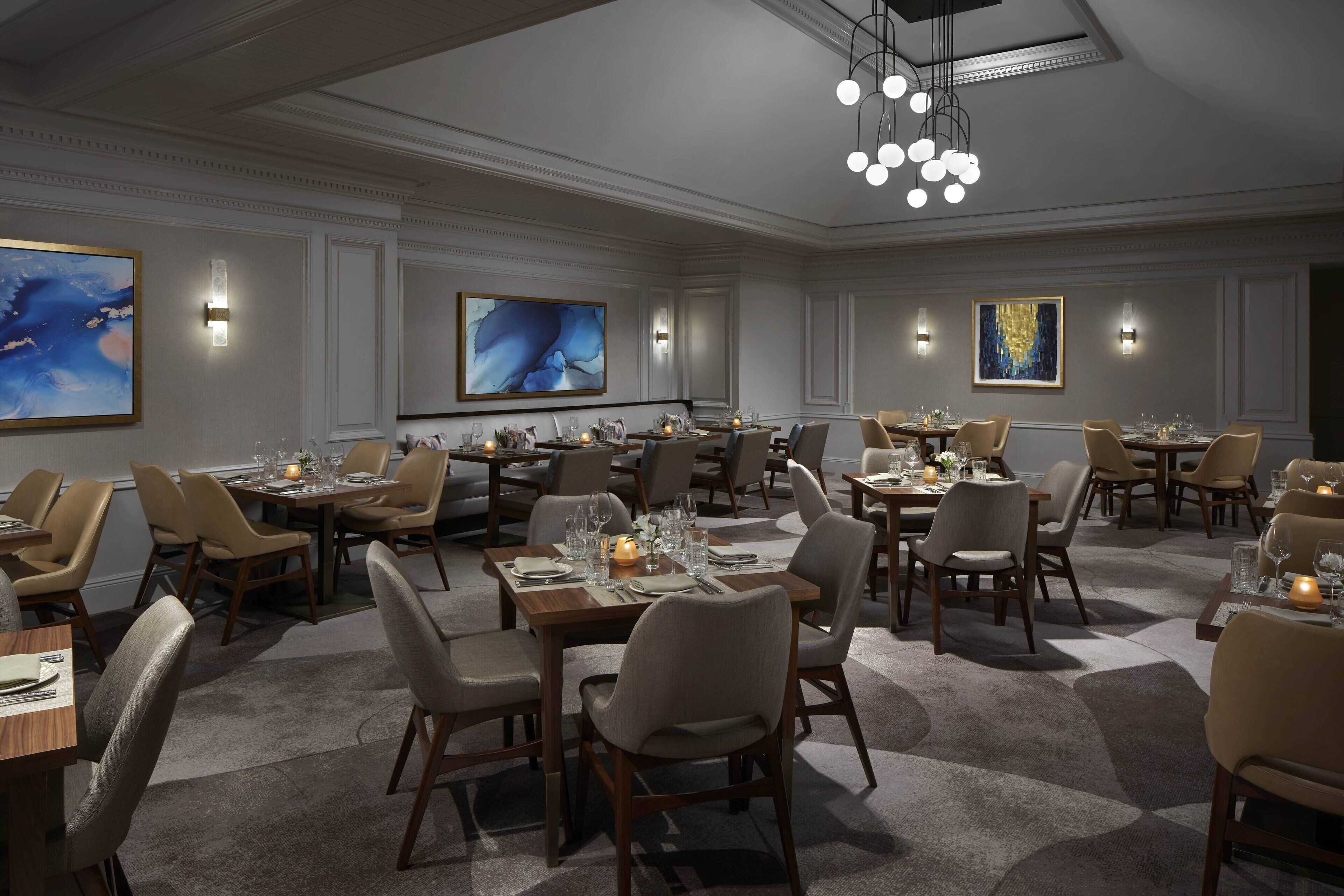 A spacious dining area with multiple tables and chairs, elegant lighting fixtures, and large abstract paintings on the walls.