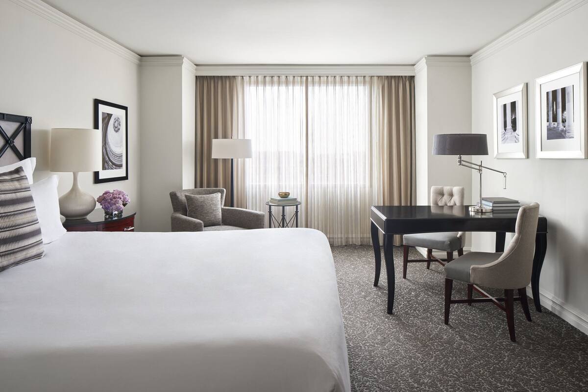 Habitación de hotel con una cama grande, un escritorio, dos sillas, una mesita y una ventana con cortinas.