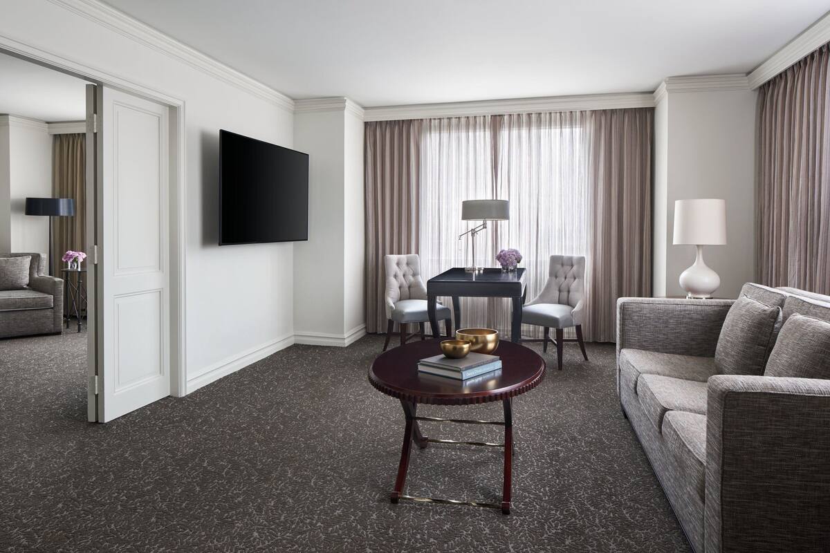 Habitación de hotel con televisor de pantalla plana, una mesa de centro redonda con libros y un cuenco, un sofá gris, dos sillones, una mesita con lámpara y un ventanal con cortinas.