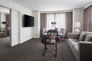 A hotel room with a flat-screen TV, a round coffee table with books and a bowl, a grey sofa, two armchairs, a small table with a lamp, and a large window with curtains.