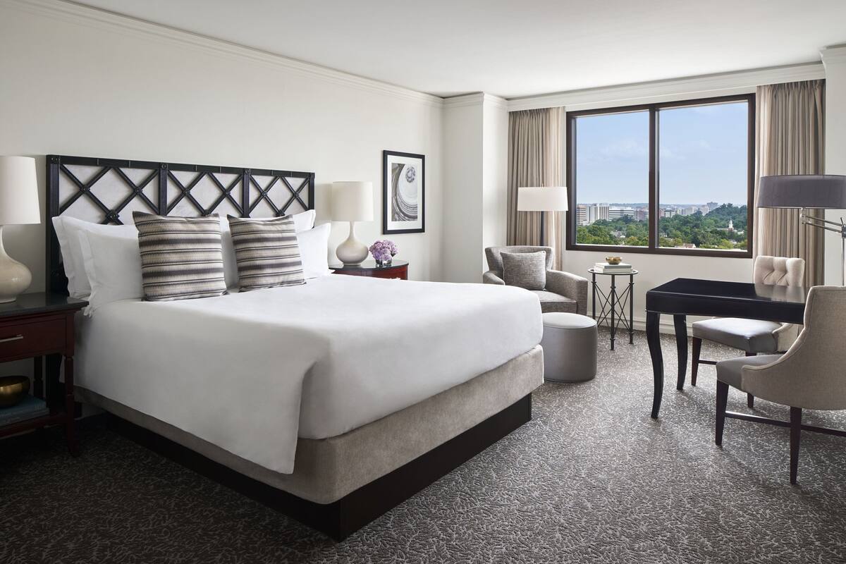Habitación de hotel con una cama grande, un escritorio, dos sillas, una mesita y vistas a la ciudad a través de amplios ventanales.