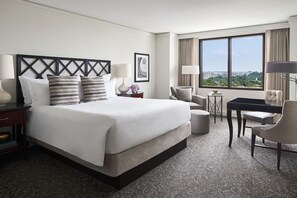 A hotel room with a large bed, a desk, two chairs, a small table, and a view of the city through large windows.