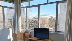 A hotel room with a bed, a desk with a TV, and large windows offering a view of buildings and trees. - La Posada de El Chaflán (Madrid)
