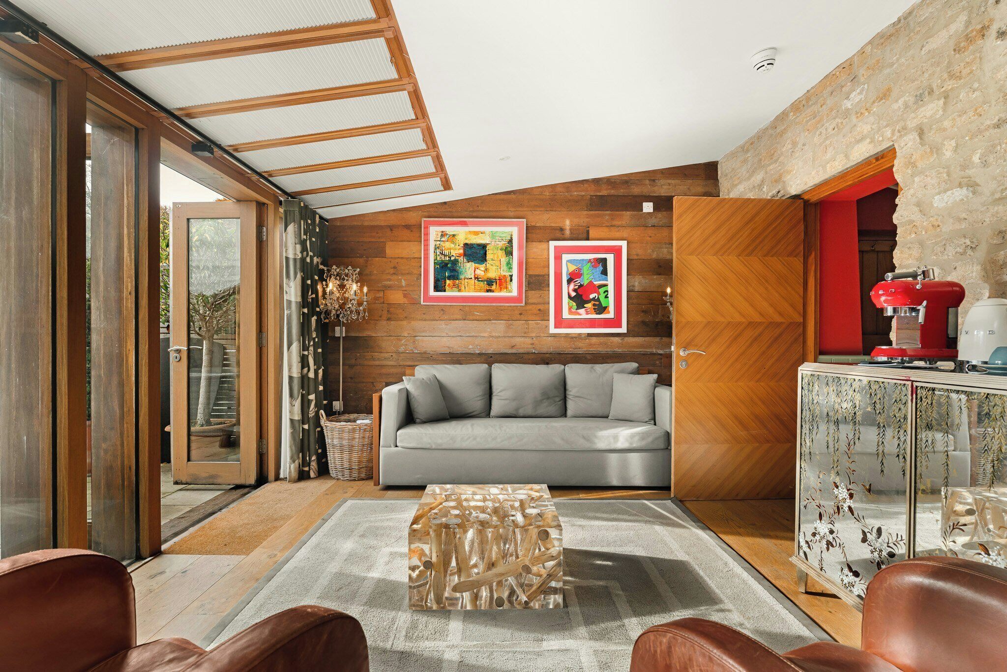 A modern living room with a grey sofa, wooden walls, and a glass door leading outside.