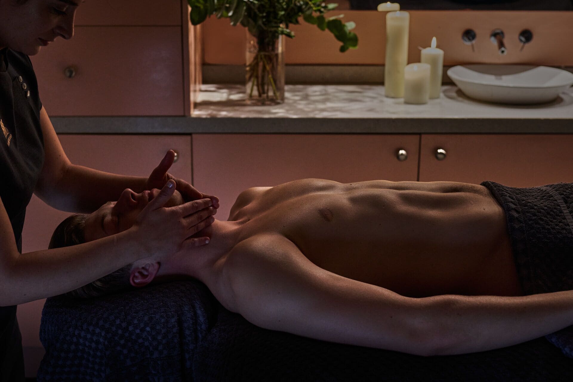 A massage therapy session in a room with a wooden cabinet, a vase with flowers, and candles on a table.