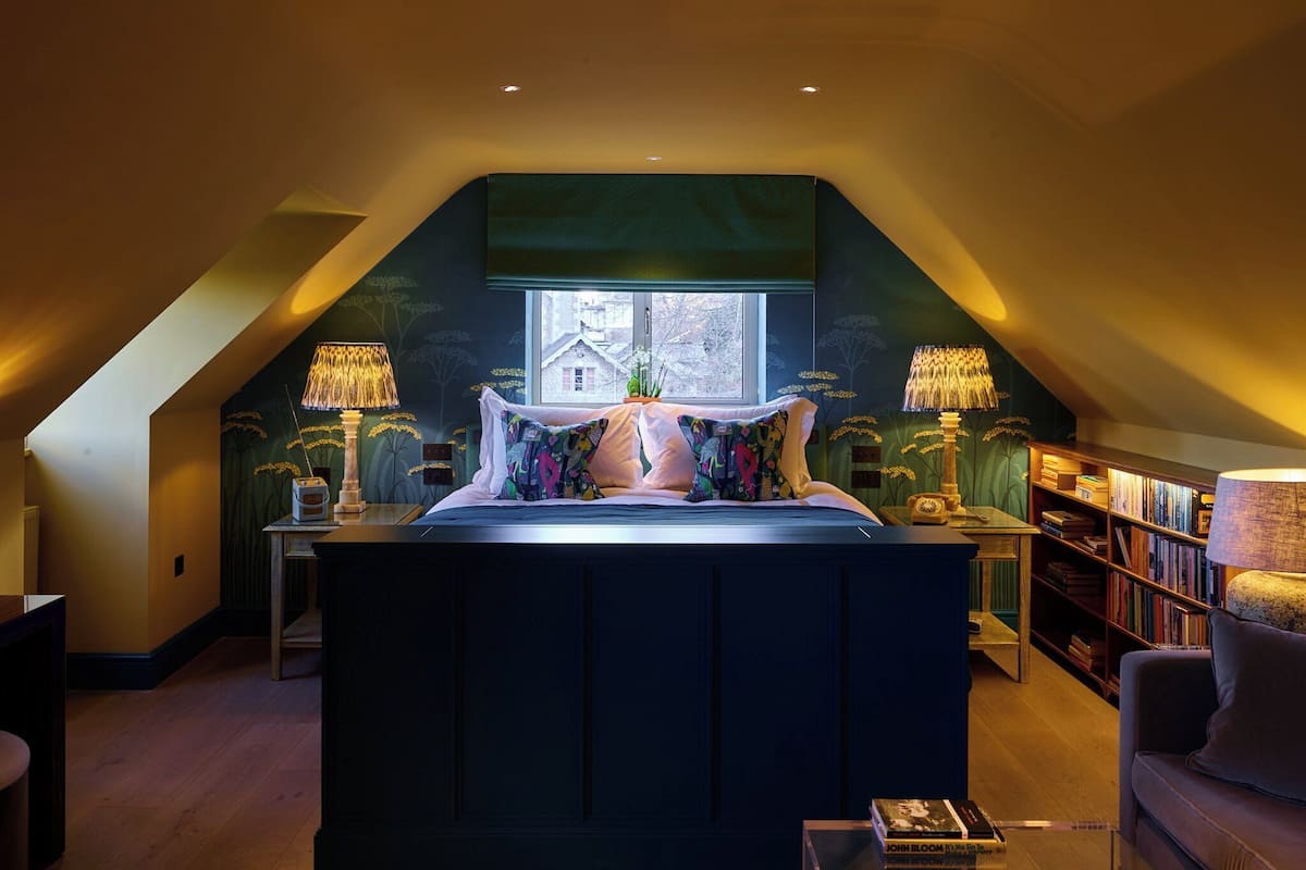 A bedroom with a sloped ceiling, a large bed, two bedside lamps, a window, and a bookshelf.