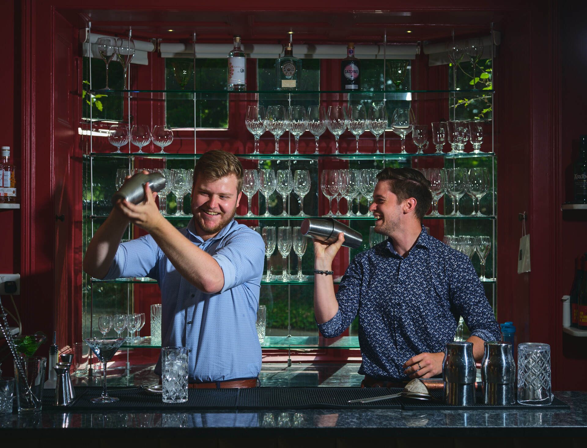 Two bartenders preparing drinks behind a bar with shelves of glasses and bottles.