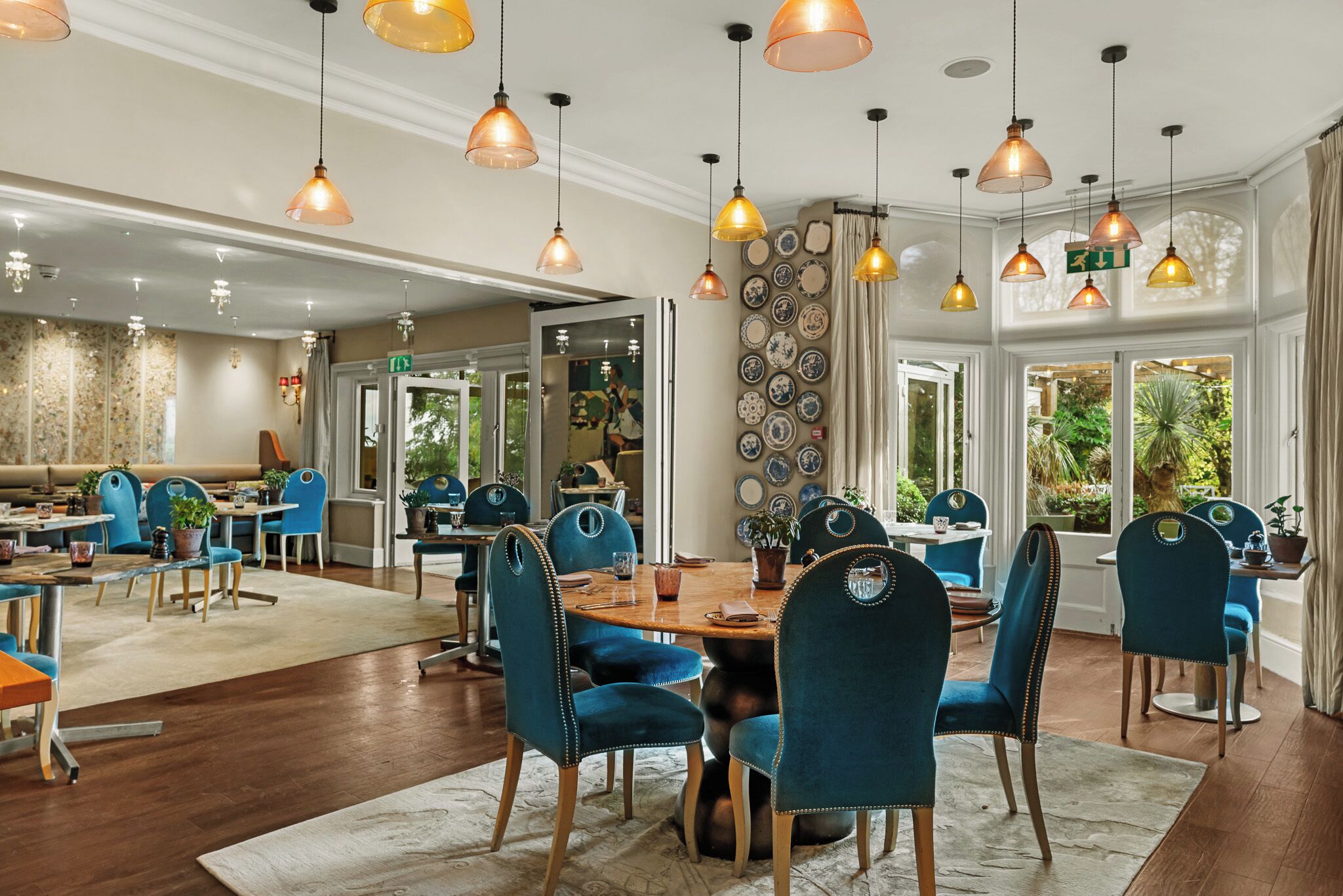 A spacious dining area with multiple tables and chairs, a decorative wall with plates, and a view of greenery outside.