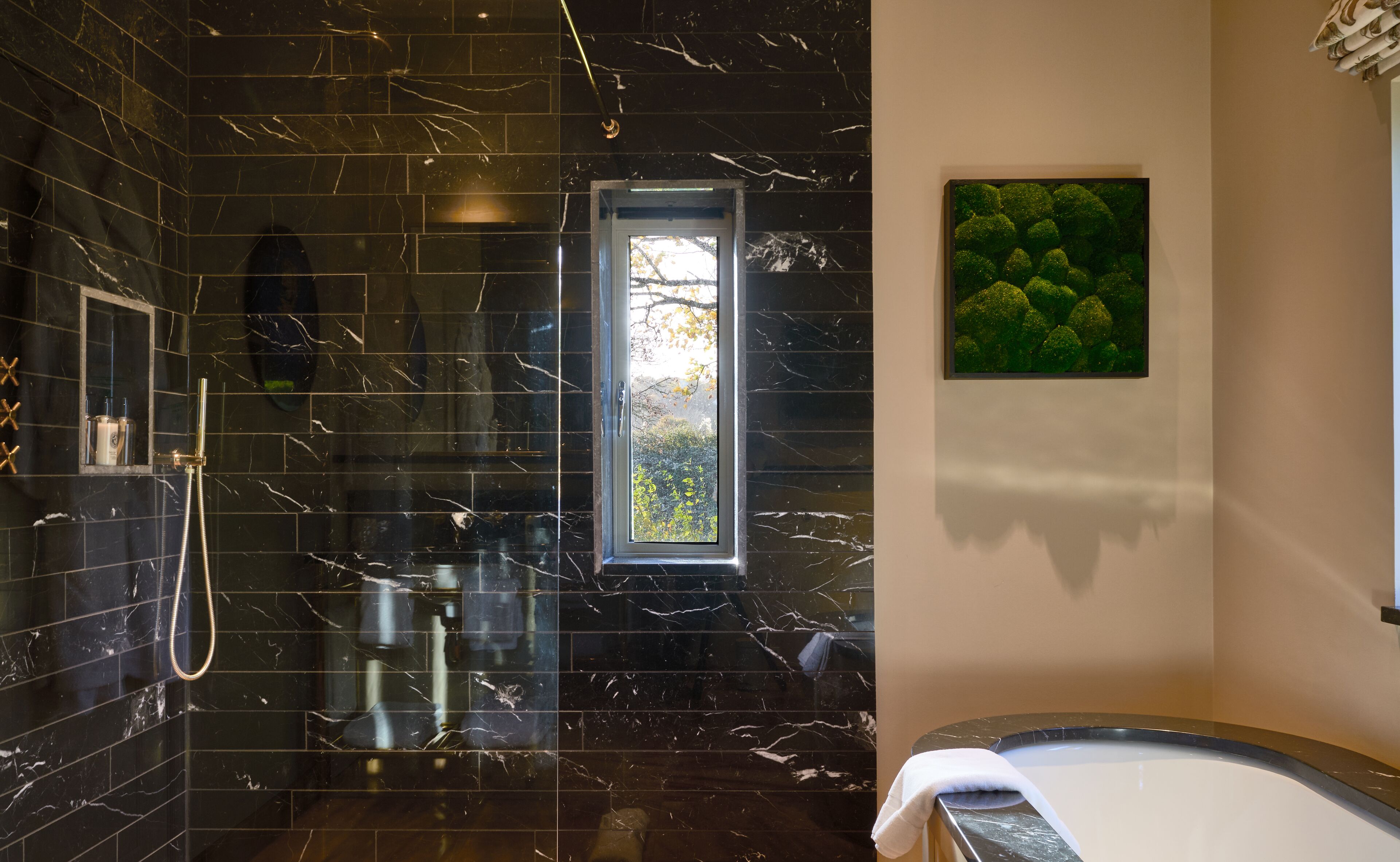 A modern bathroom with a black marble floor, a bathtub, a shower area, and a window with a view of trees.
