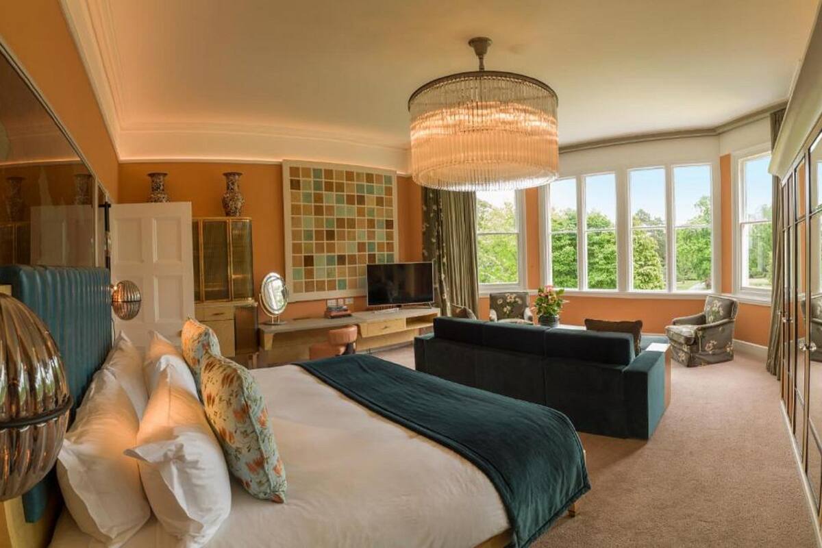 A spacious bedroom with a large bed, a seating area, a television, and a view of greenery through the windows.
