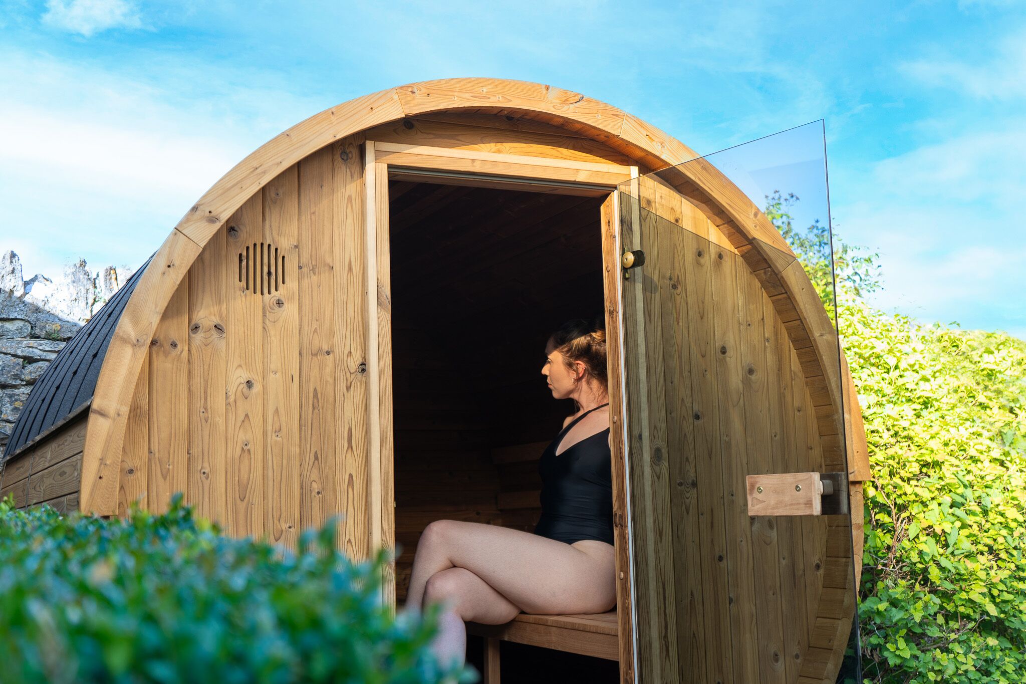 Una sauna de madera con techo curvo, una puerta y vistas del entorno cubierto de vegetación.