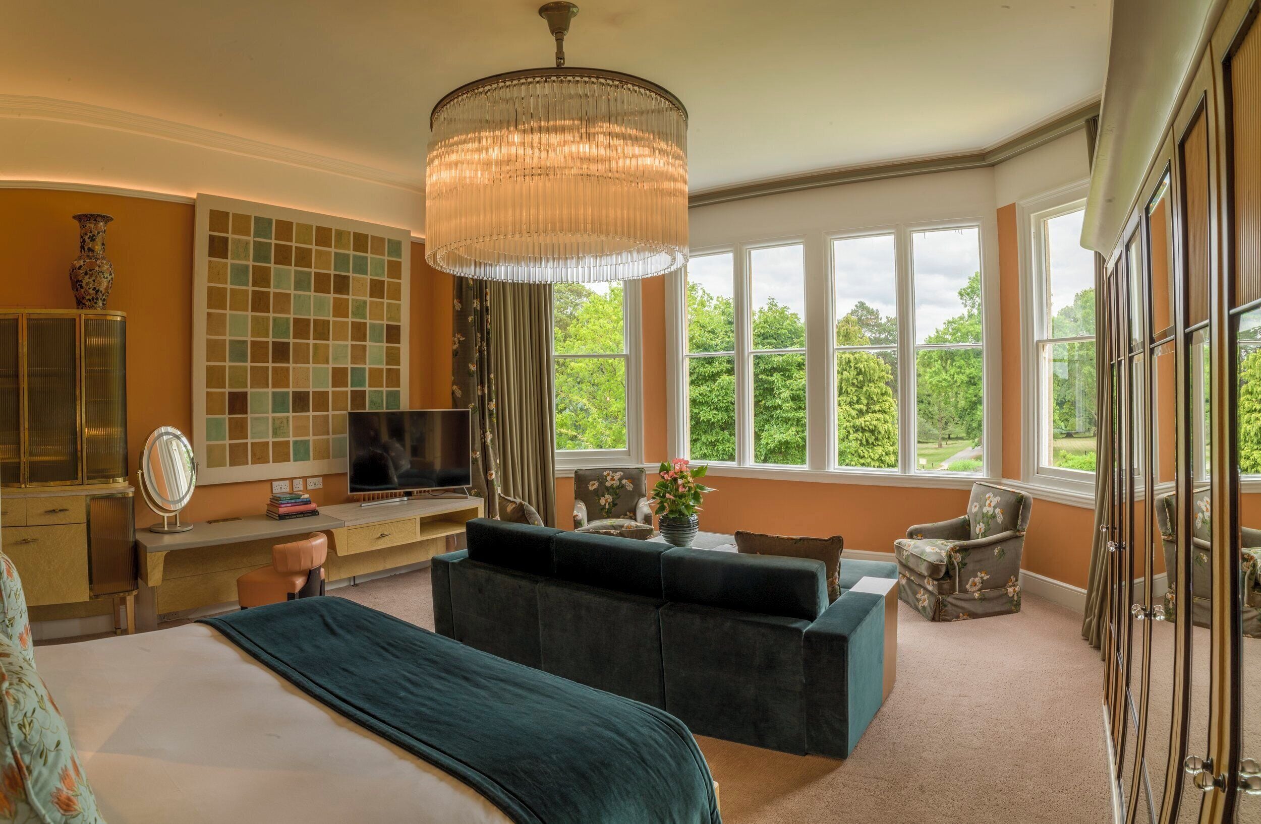 A bedroom with a large bed, a desk, a television, and a view of greenery through large windows.