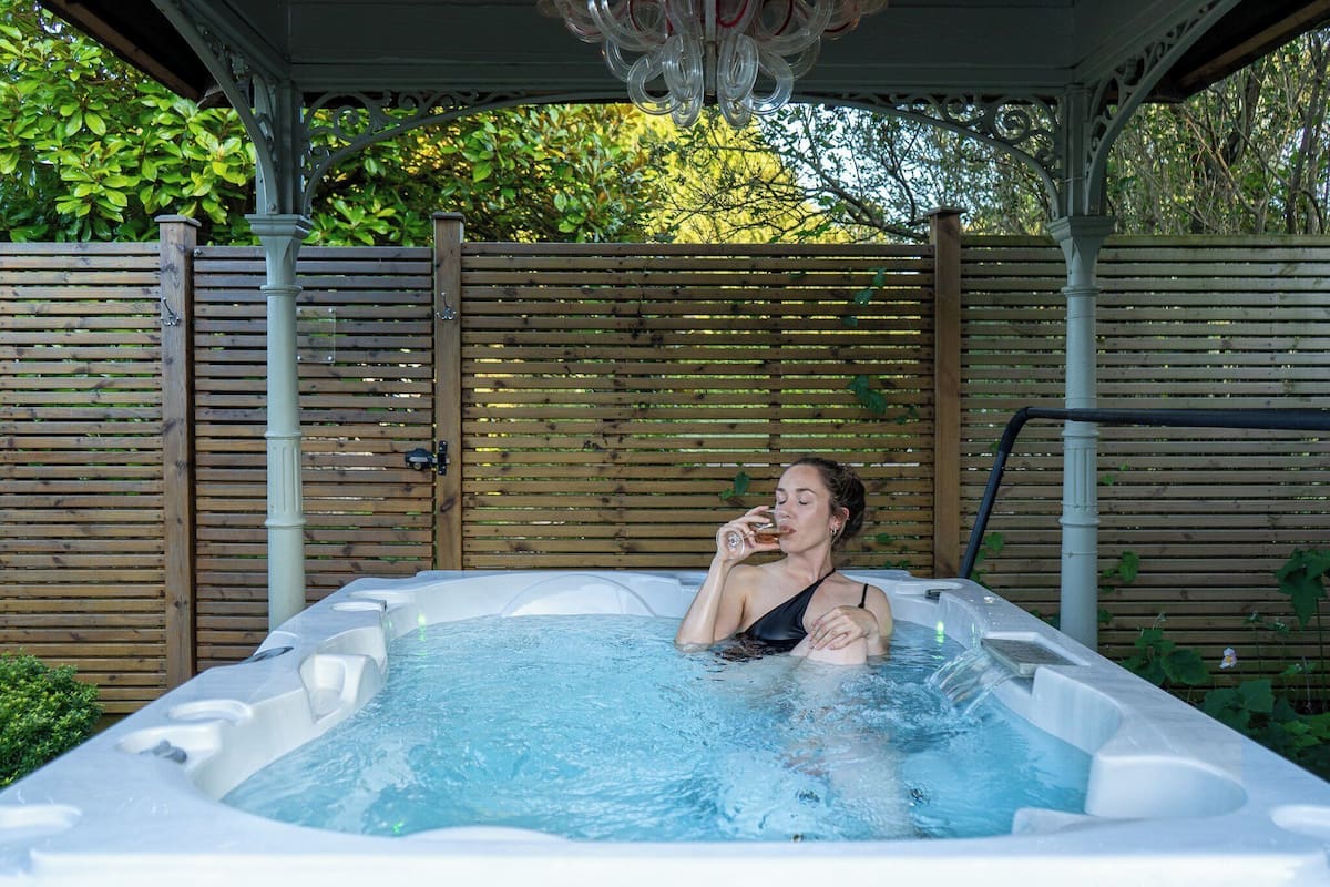 A person relaxing in a hot tub with a wooden fence and greenery in the background.
