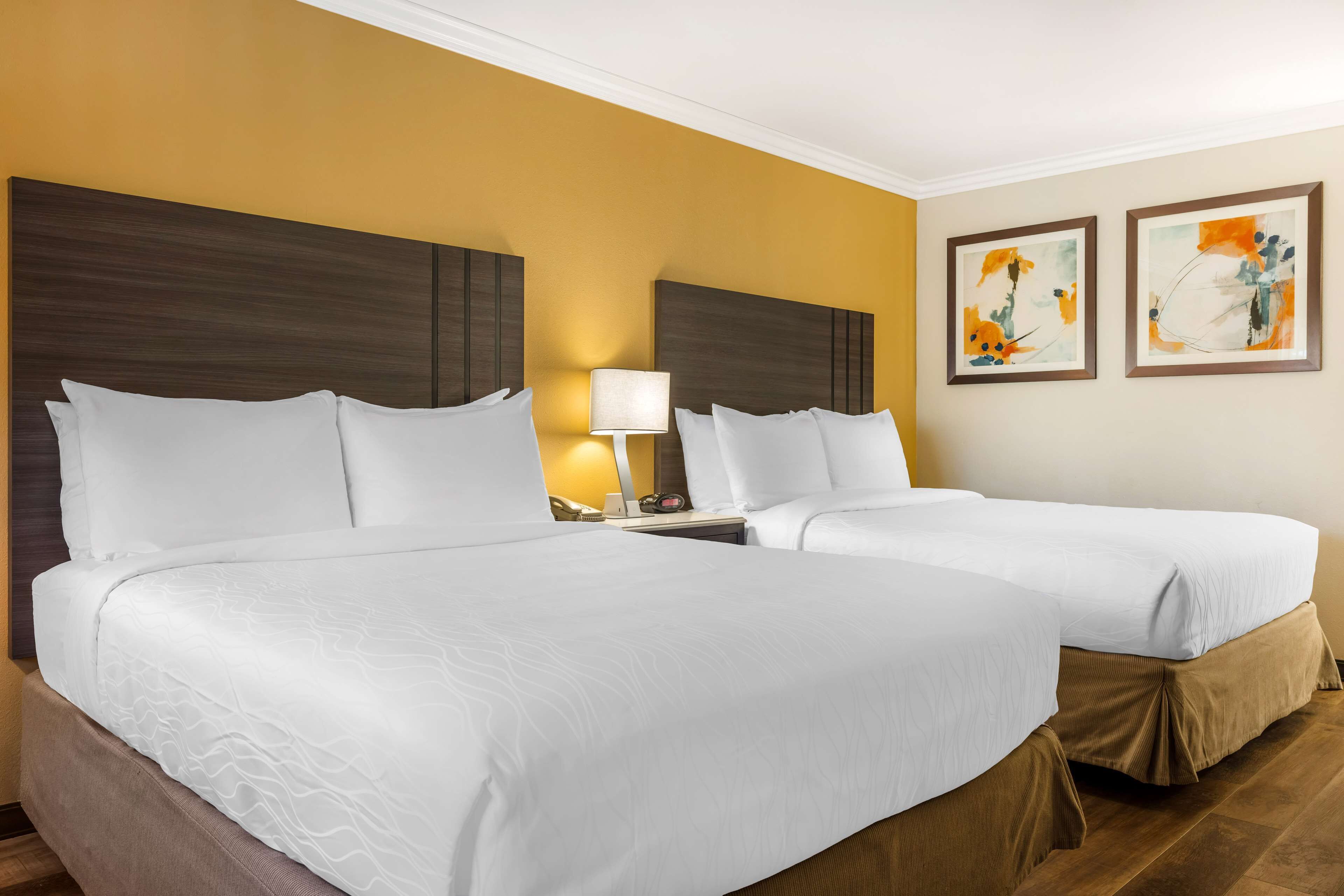 A hotel room with two beds, a nightstand with a lamp, and two framed artworks on the wall.