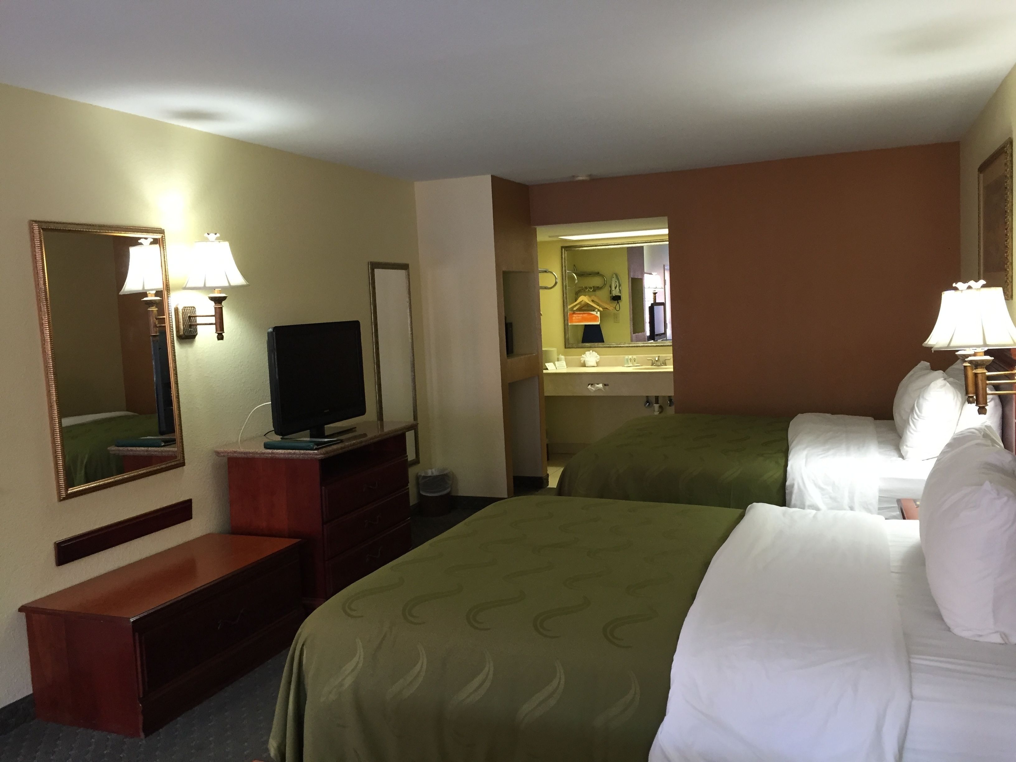 A hotel room with two beds, a TV, a dresser, a mirror, and a bathroom visible through an open door.