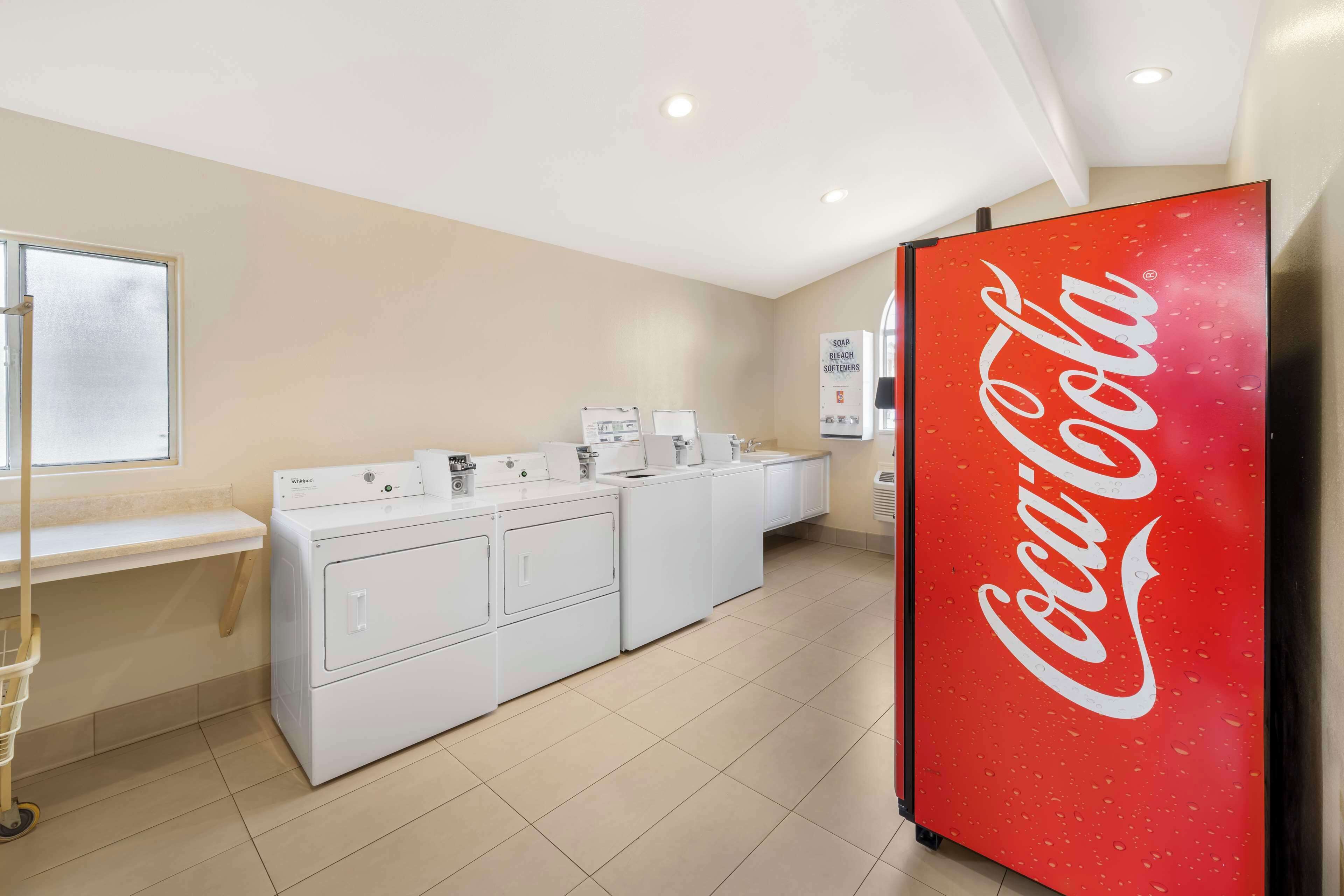 Ein Waschbereich mit mehreren Waschmaschinen, einem Coca-Cola-Kühlschrank und einem Fenster mit Jalousien.