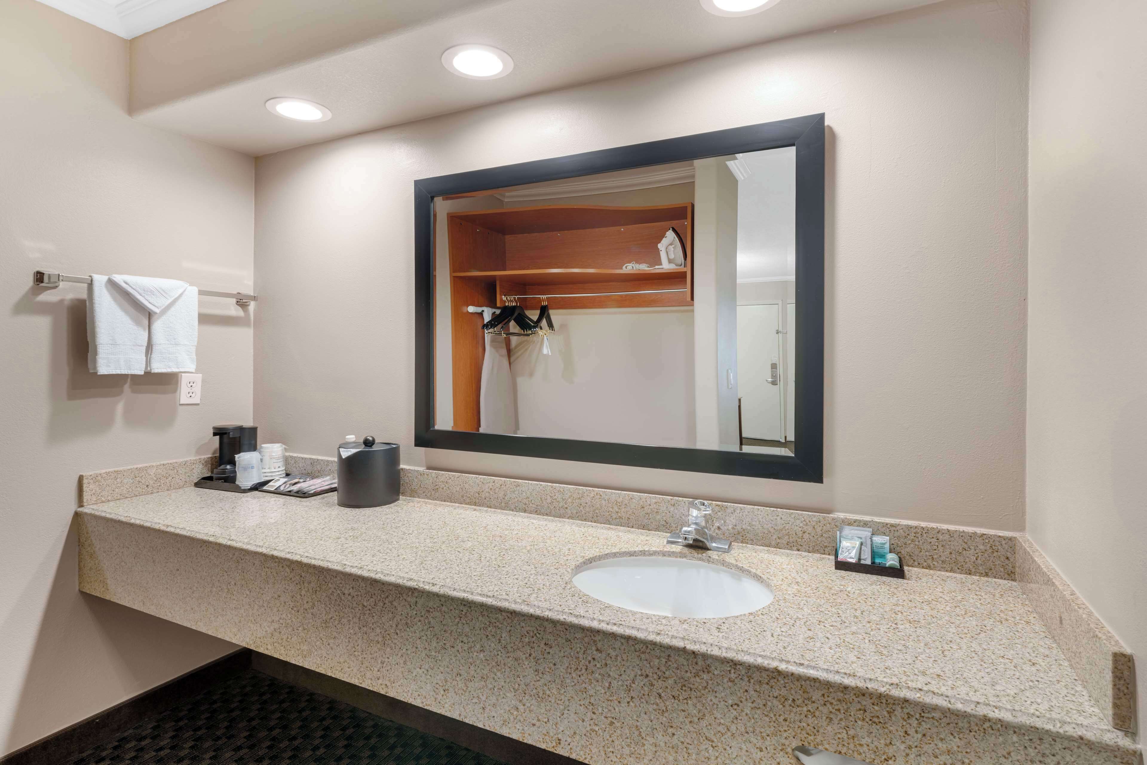 Un baño de hotel con encimera de granito, lavabo, espejo, toallero y estante con artículos de aseo.