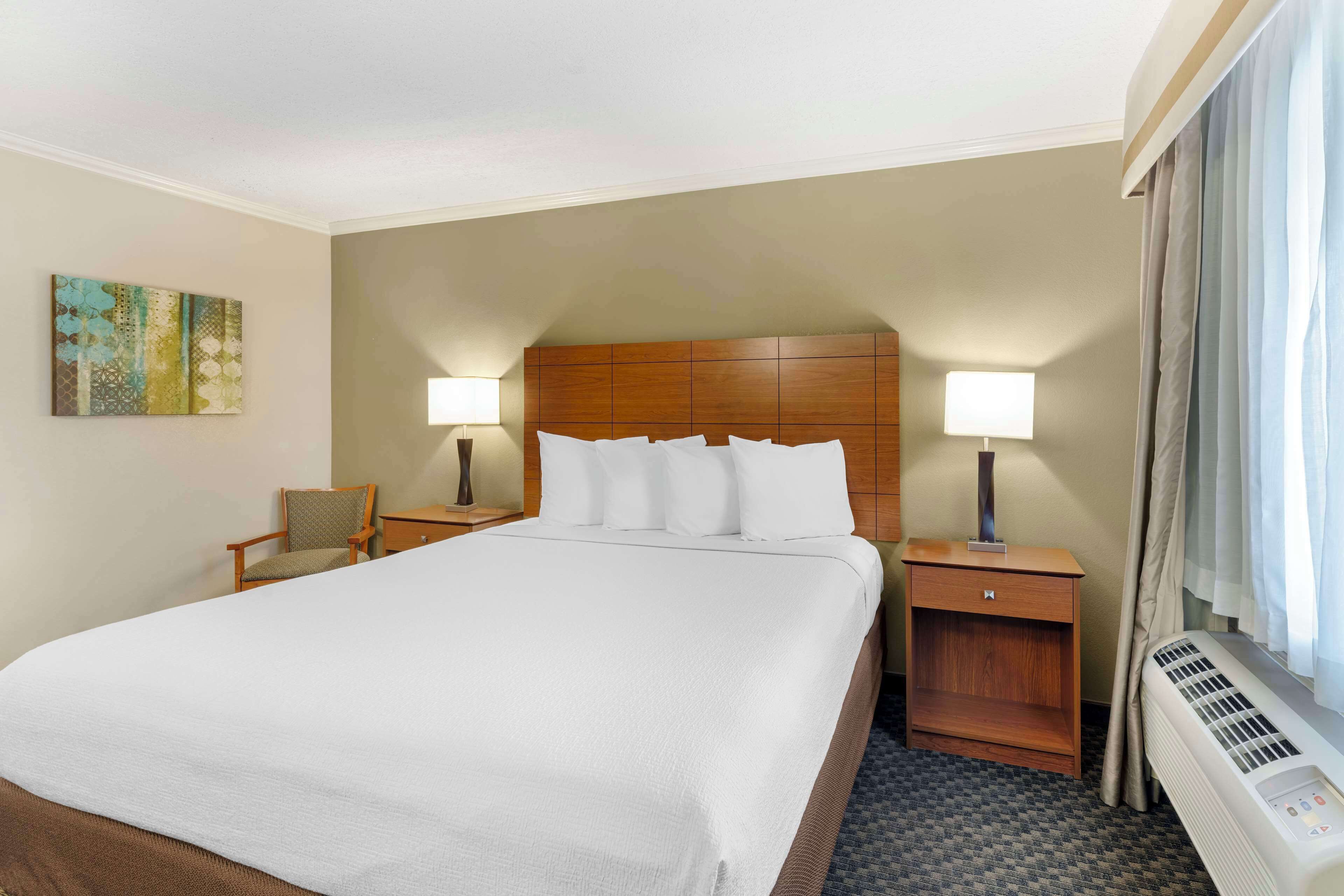 A hotel room with a large bed, two bedside tables with lamps, a chair, a small cabinet, and a window with curtains.