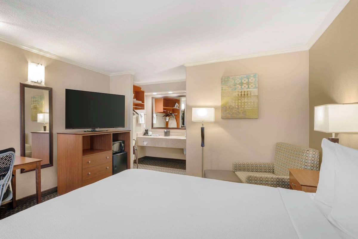 A hotel room with a large bed, a television, a desk with a chair, a small table, a lamp, a mirror, and a wardrobe.