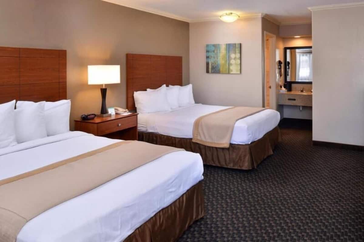 A hotel room with two beds, a nightstand with a lamp, a painting on the wall, and a bathroom visible through an open door.