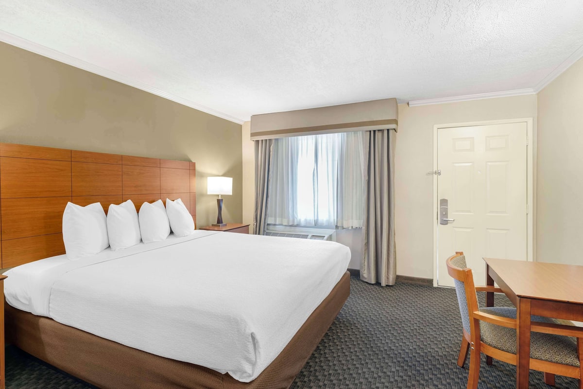 A hotel room with a large bed, a wooden headboard, a desk with a chair, a window with curtains, and a door.