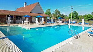 A swimming pool with lounge chairs and umbrellas, a brick building with a red roof, and a clear blue sky. - Days Inn & Suites by Wyndham Williamsburg Colonial (Williamsburg)
