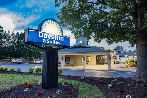 A Days Inn & Suites sign with a sunburst design, a building entrance, and a landscaped area with flowers. - Days Inn & Suites by Wyndham Williamsburg Colonial (Williamsburg)