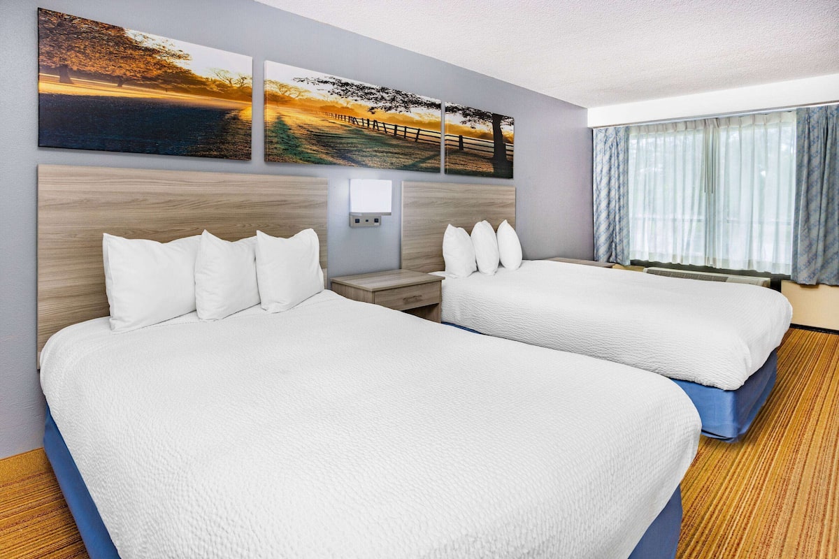 A hotel room with two beds, a headboard, a nightstand, and a window with curtains.