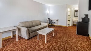 A compact hotel room with a sofa, a small desk, a chair, and a compact kitchenette. - Days Inn & Suites by Wyndham Williamsburg Colonial (Williamsburg)
