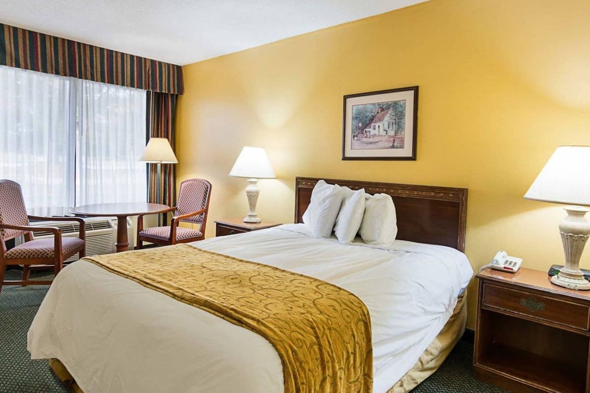 A hotel room with a bed, two chairs, a table, two lamps, a painting on the wall, and a telephone on the bedside table.
