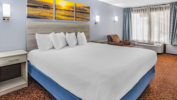 A hotel room with a large bed, a nightstand, a chair, and a window with curtains. - Days Inn & Suites by Wyndham Williamsburg Colonial (Williamsburg)