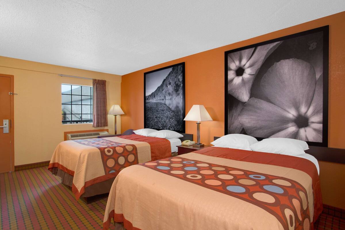 Ein Hotelzimmer mit zwei Betten, einem Fenster mit Vorhängen, einem an der Wand montierten Fernseher und einem großen Blumenbild.