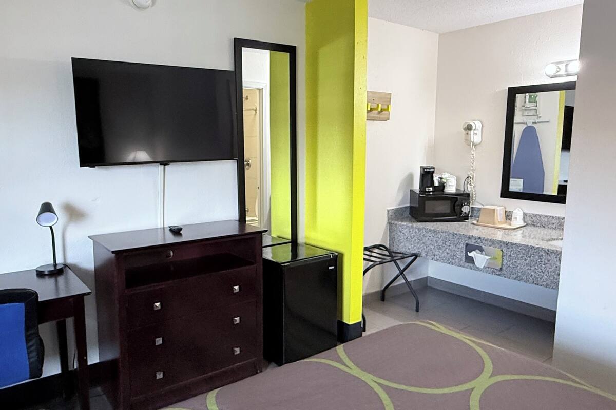 Ein Hotelzimmer mit Fernseher, Schreibtisch, Kommode, Stuhl, Spiegel und einem Badezimmer mit Waschbecken und Mikrowelle.