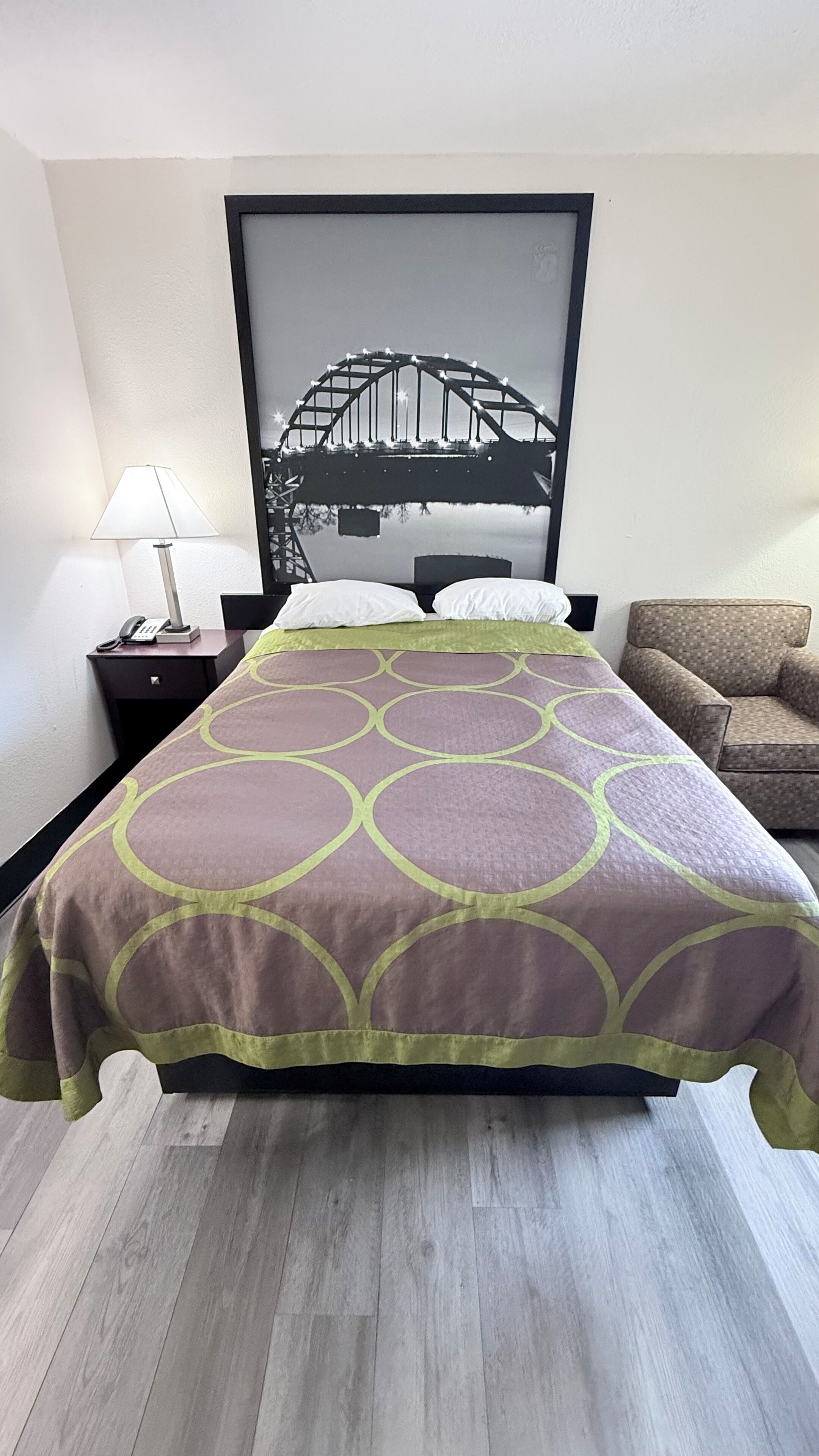 A hotel room with a bed, a nightstand with a lamp, a framed picture of a bridge, and a sofa.