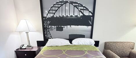 A hotel room with a bed, a nightstand with a lamp, a framed picture of a bridge, and a sofa.