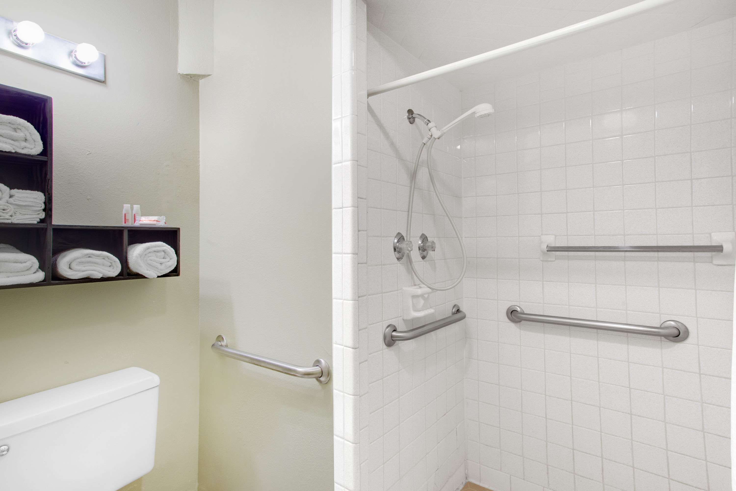 A bathroom with a toilet, a towel rack with folded towels, a shower with grab bars, and a mirror with lights above it.