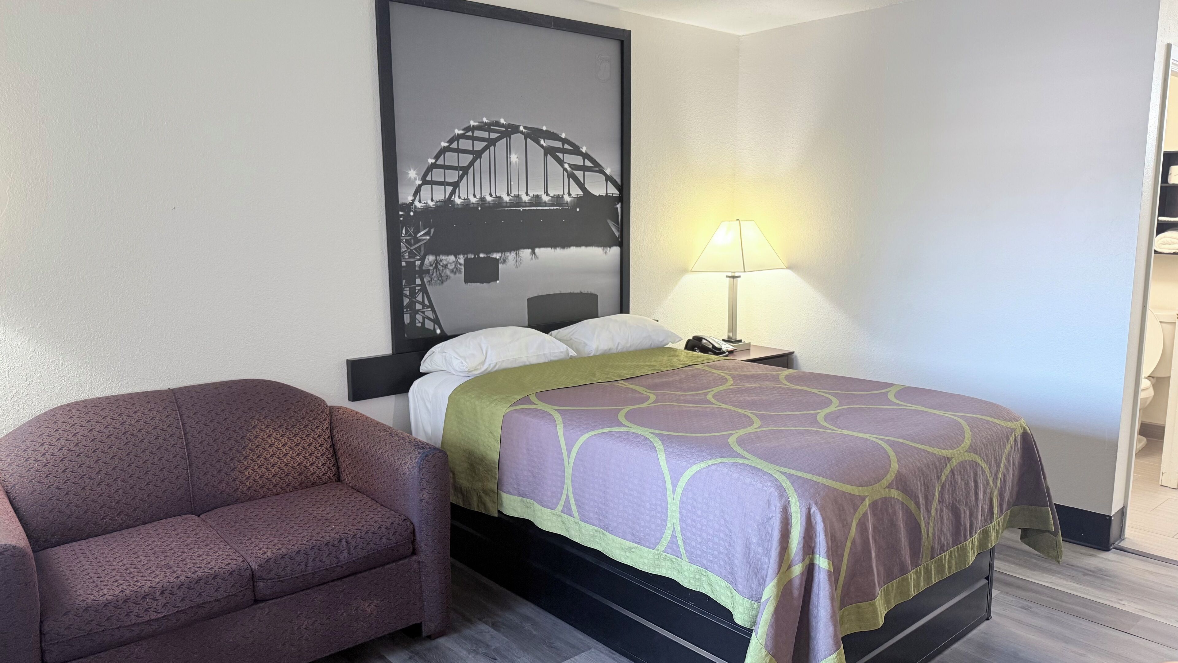 A hotel room with a bed, a chair, a nightstand with a lamp, and a large framed picture of a bridge.