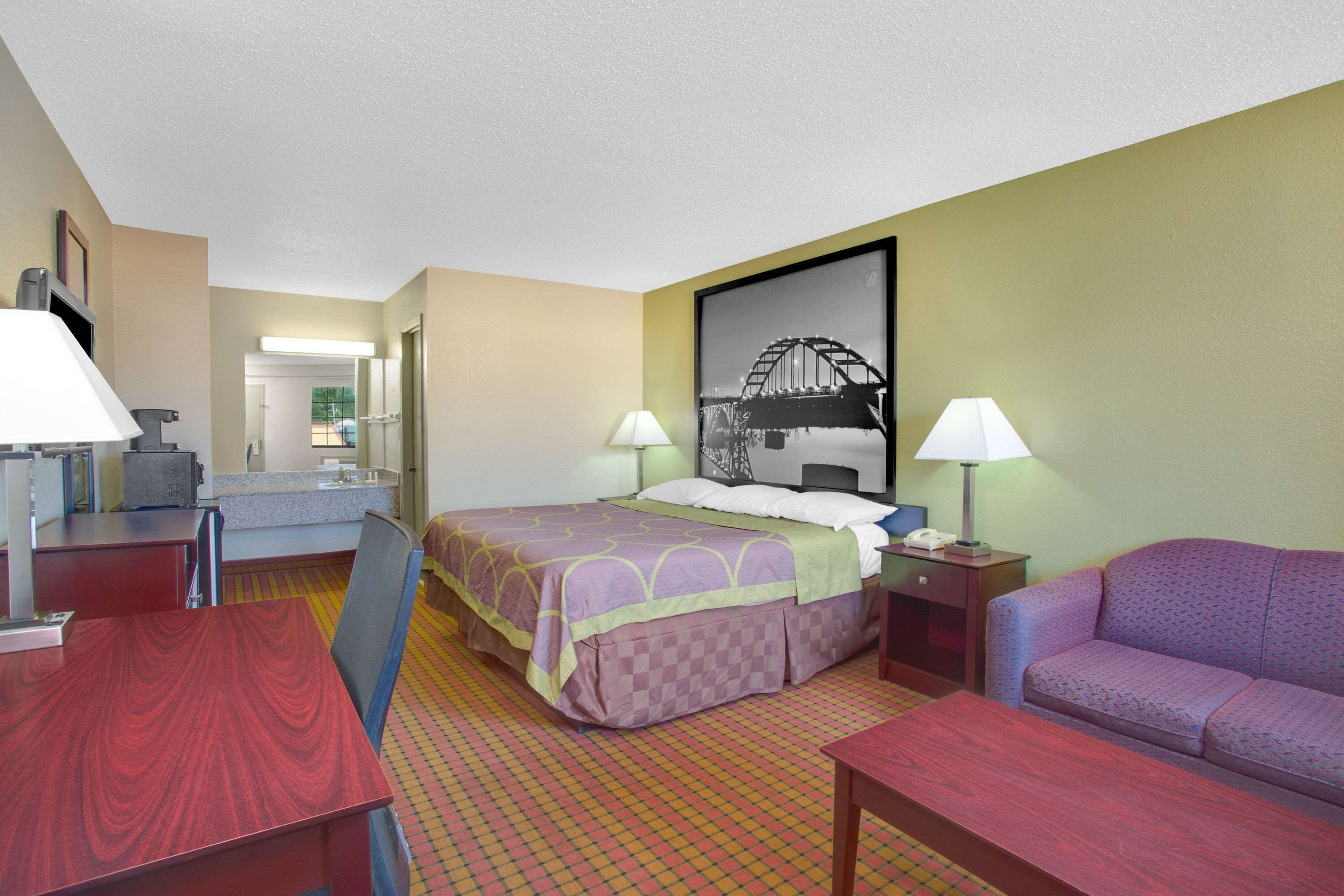 A hotel room with a bed, a desk, a chair, a sofa, and a bathroom visible in the background.