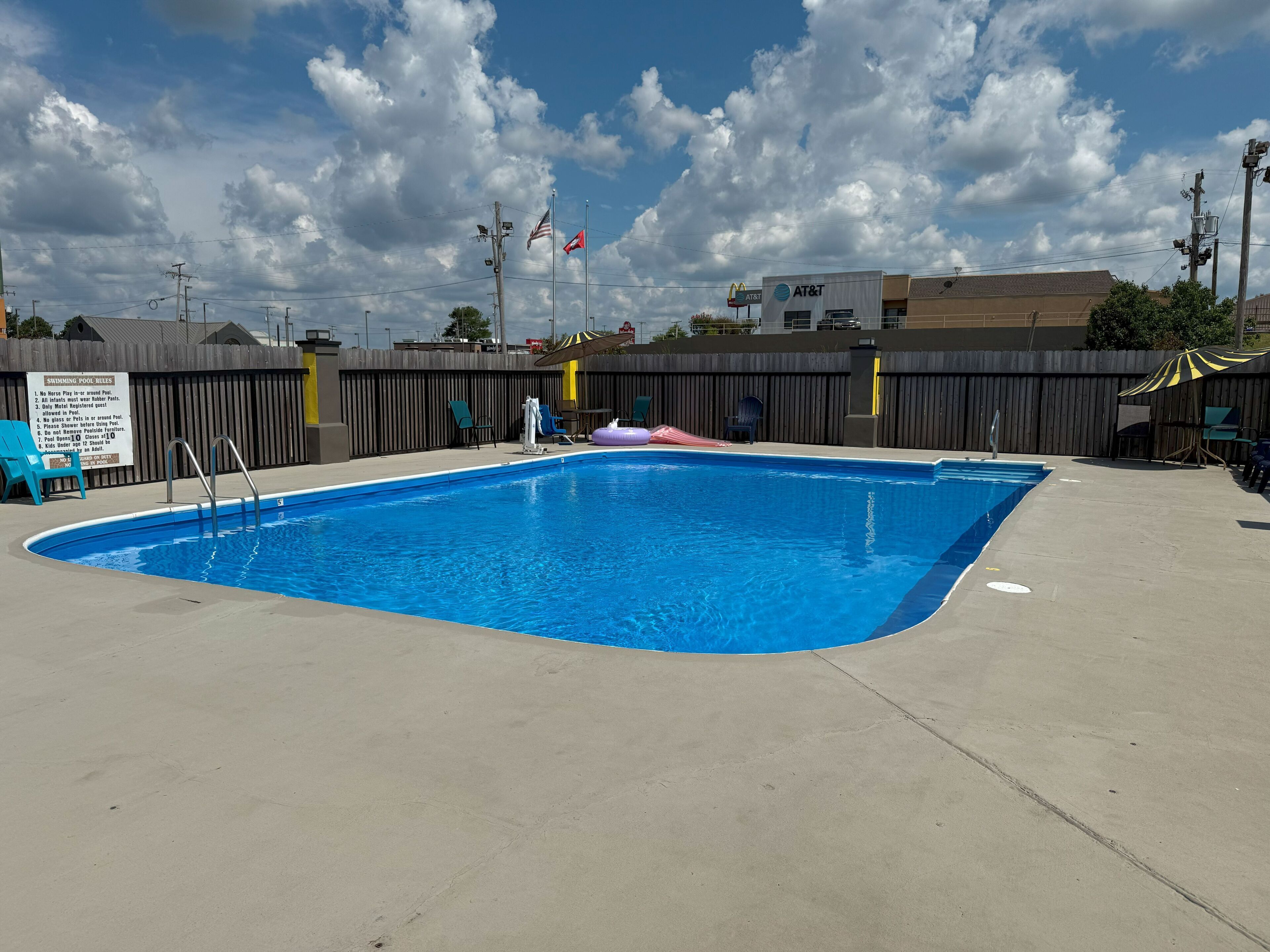Una piscina con zona vallada, sillas de descanso y un cartel con las normas de la piscina.