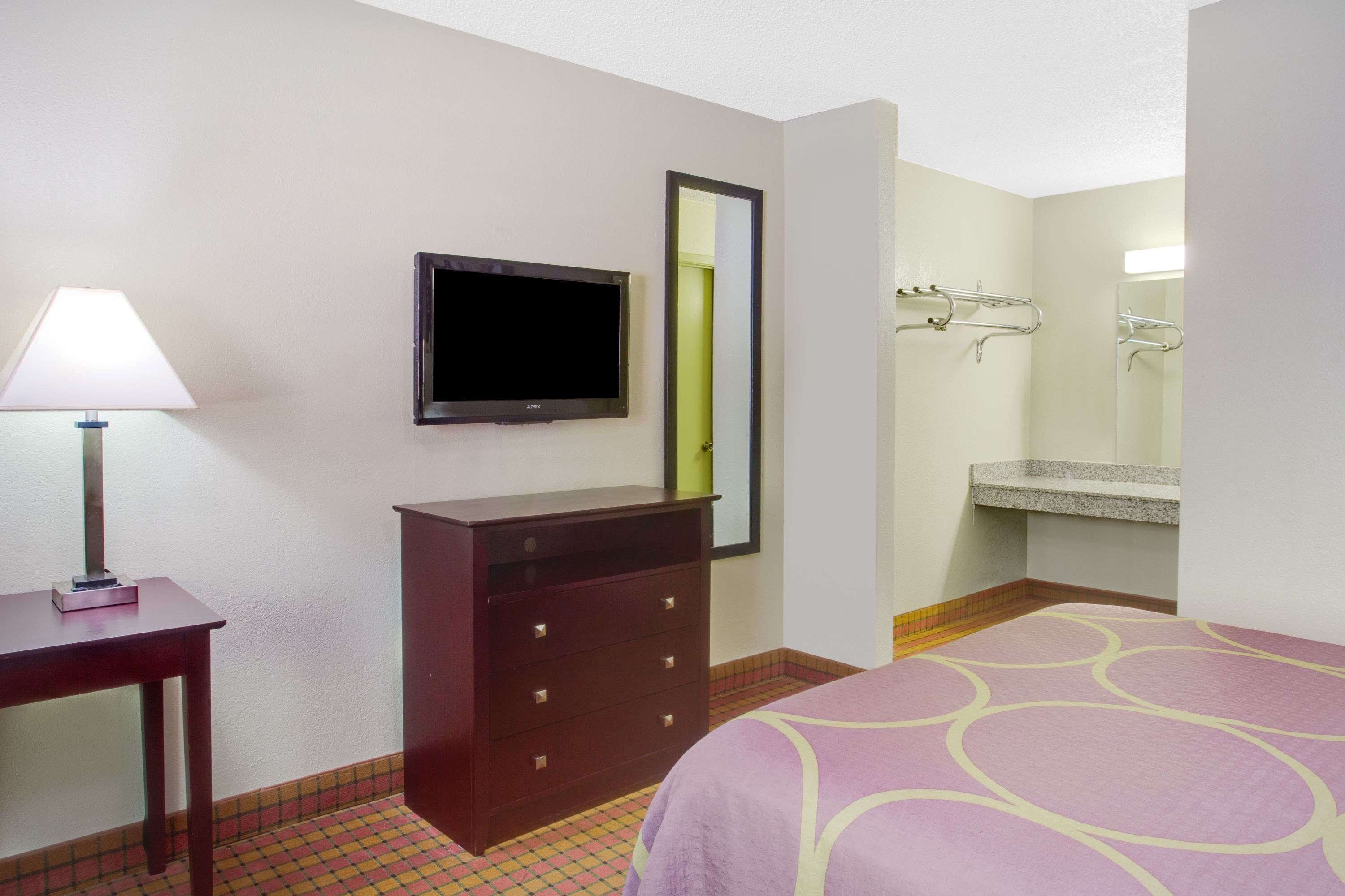 Habitación de hotel con cama, cómoda, televisor, espejo y baño con lavamanos y perchero para toallas.