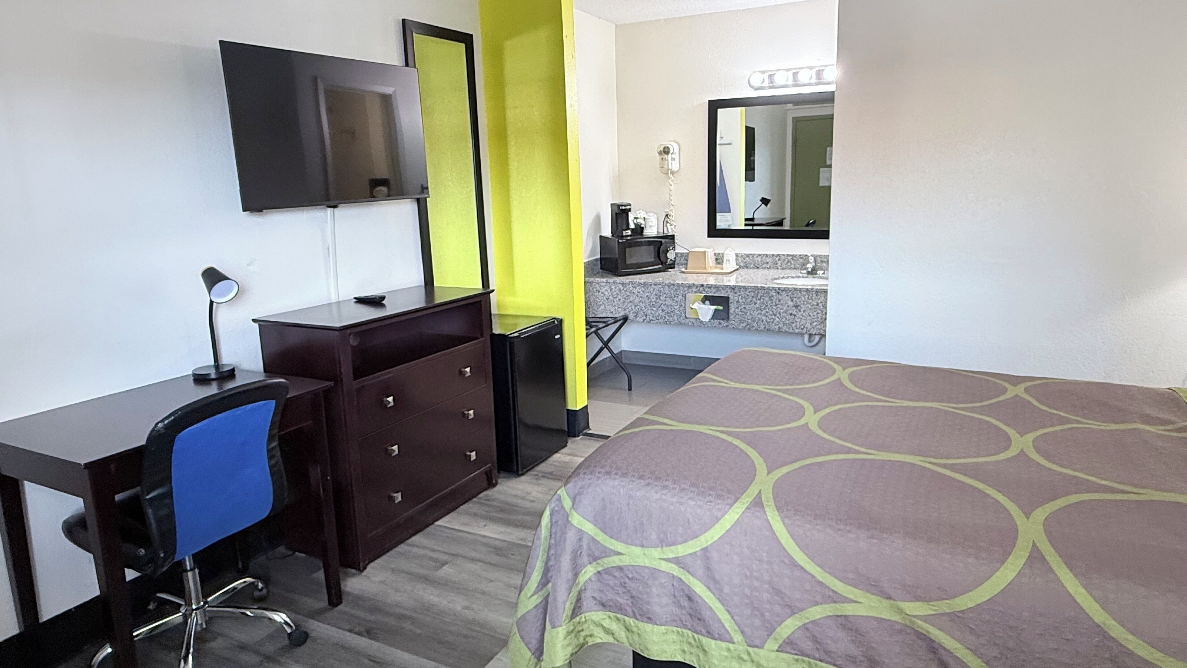 A hotel room with a bed, desk, chair, dresser, and a bathroom with a sink and mirror.
