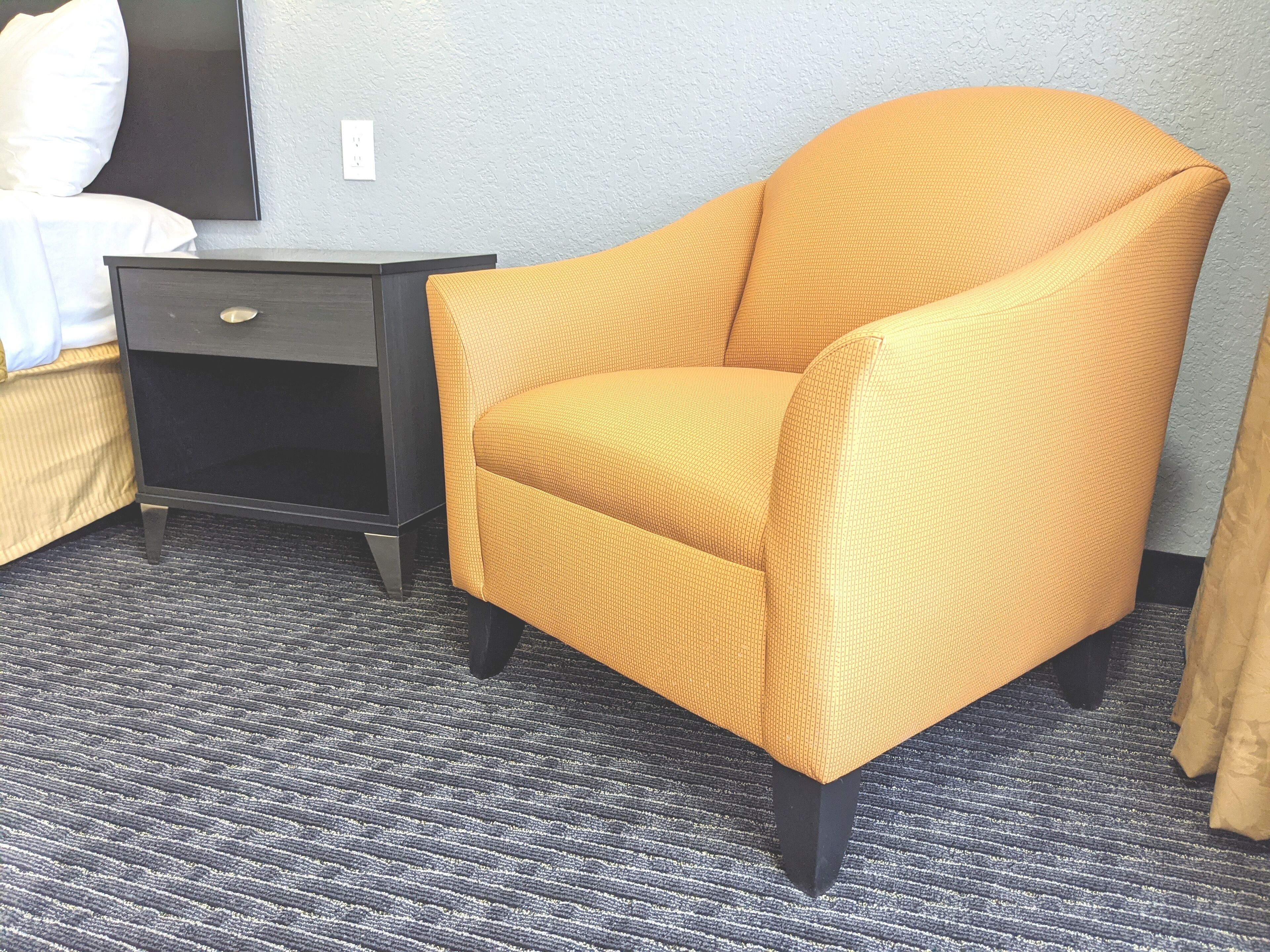 A room with a yellow armchair, a dark nightstand with a drawer, and a bed with white bedding.