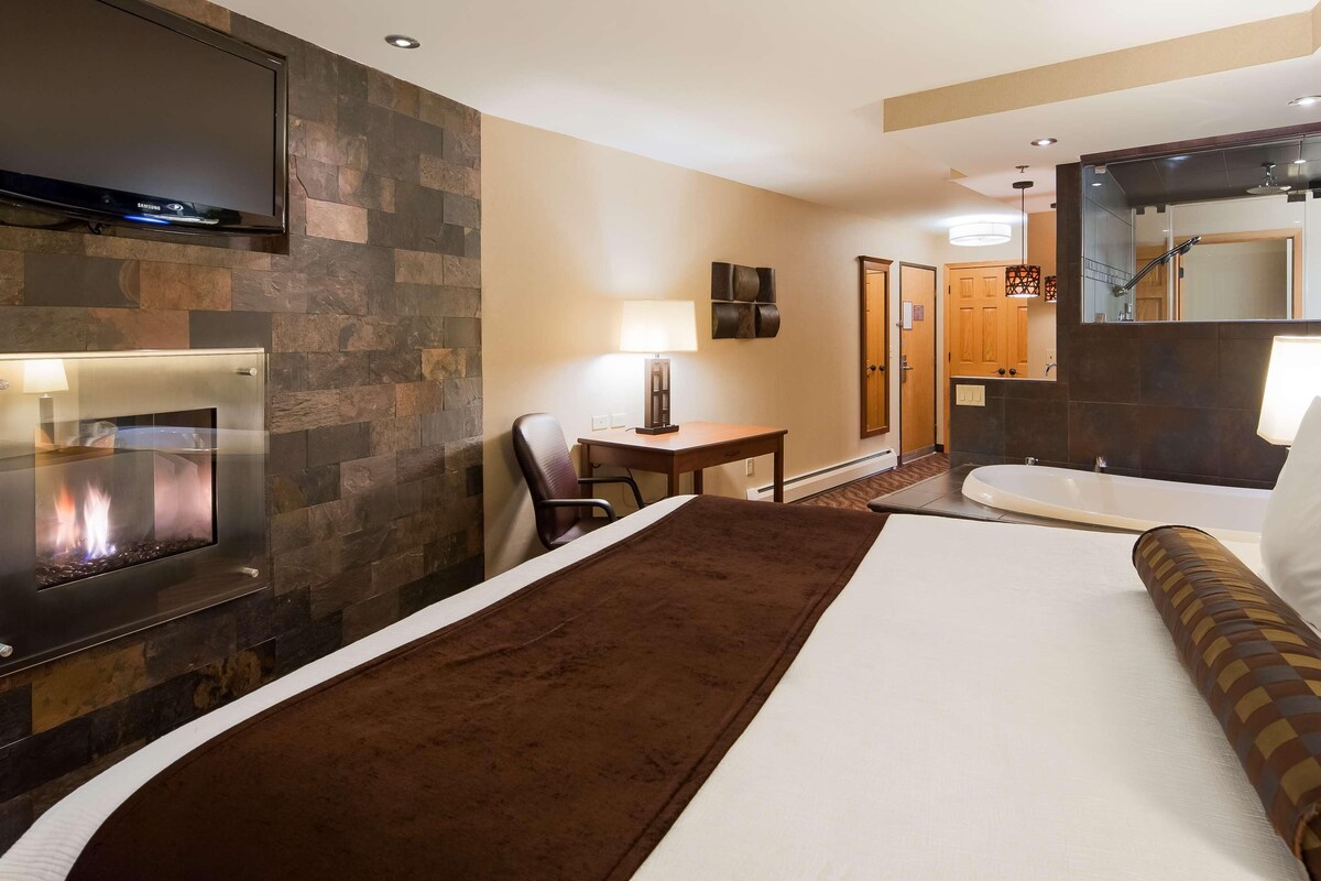 Quarto de hotel com cama, escrivaninha, cadeira, televisão, lareira e banheiro com banheira e espelho.