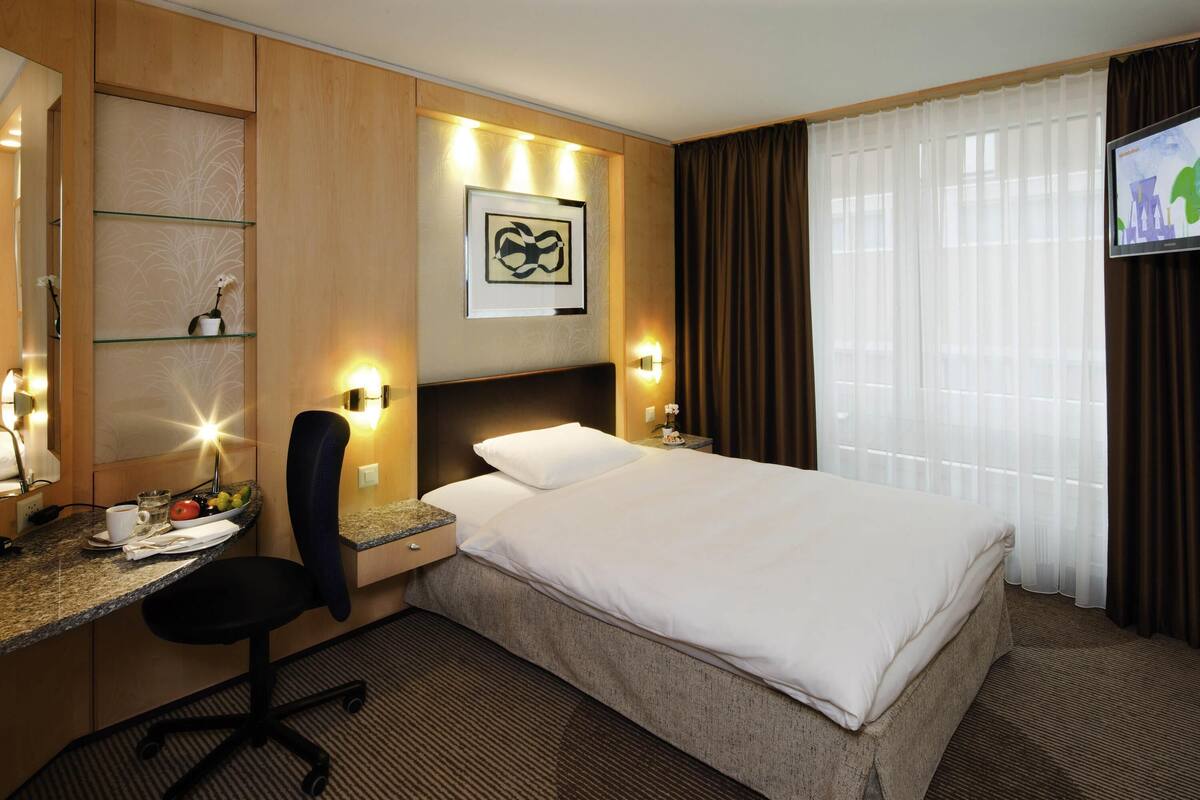 Une chambre d’hôtel avec un lit, un bureau et une chaise, une télévision et une fenêtre avec des rideaux.
