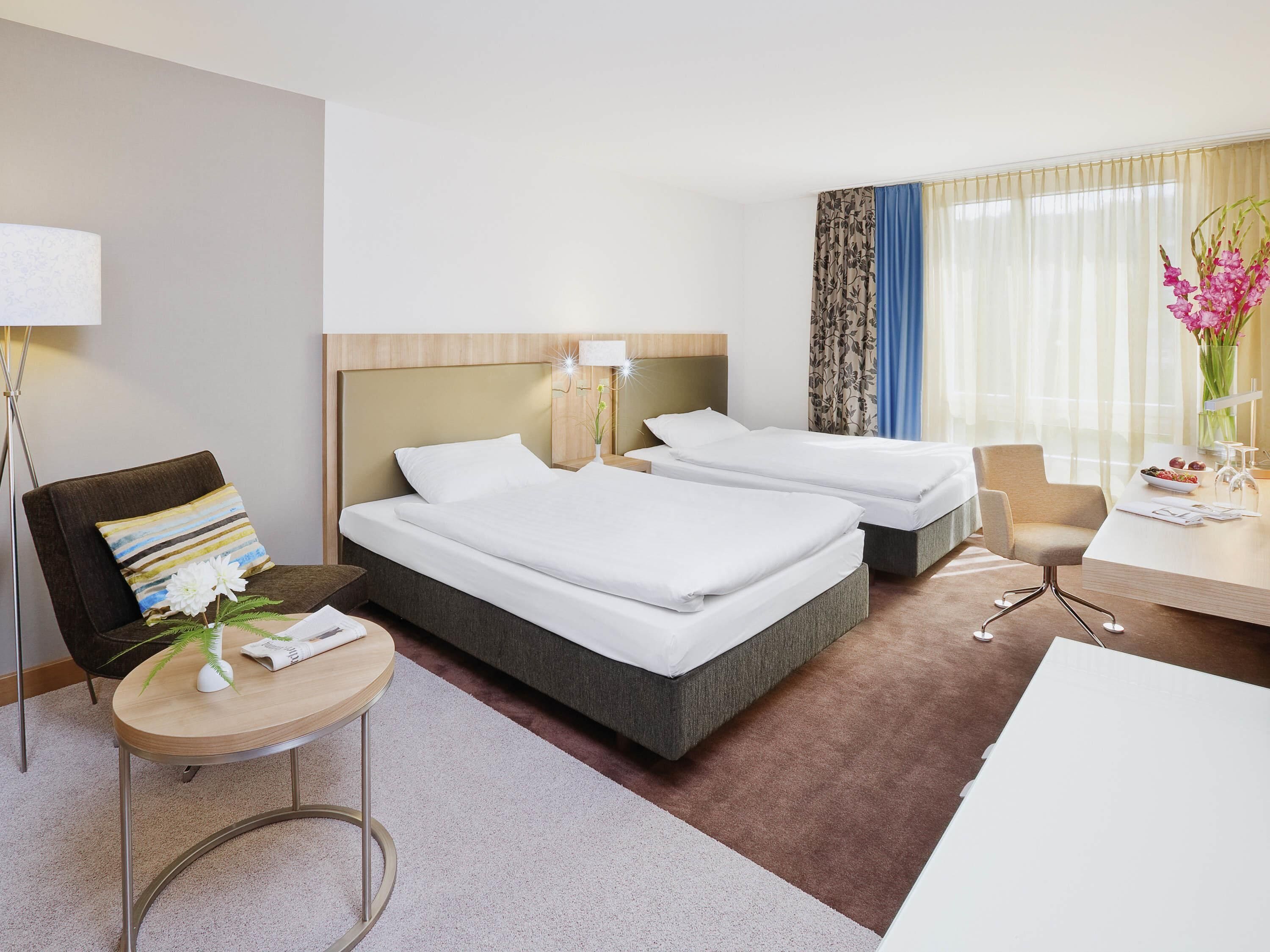 A hotel room with two beds, a desk, a chair, a lamp, and a vase with flowers.