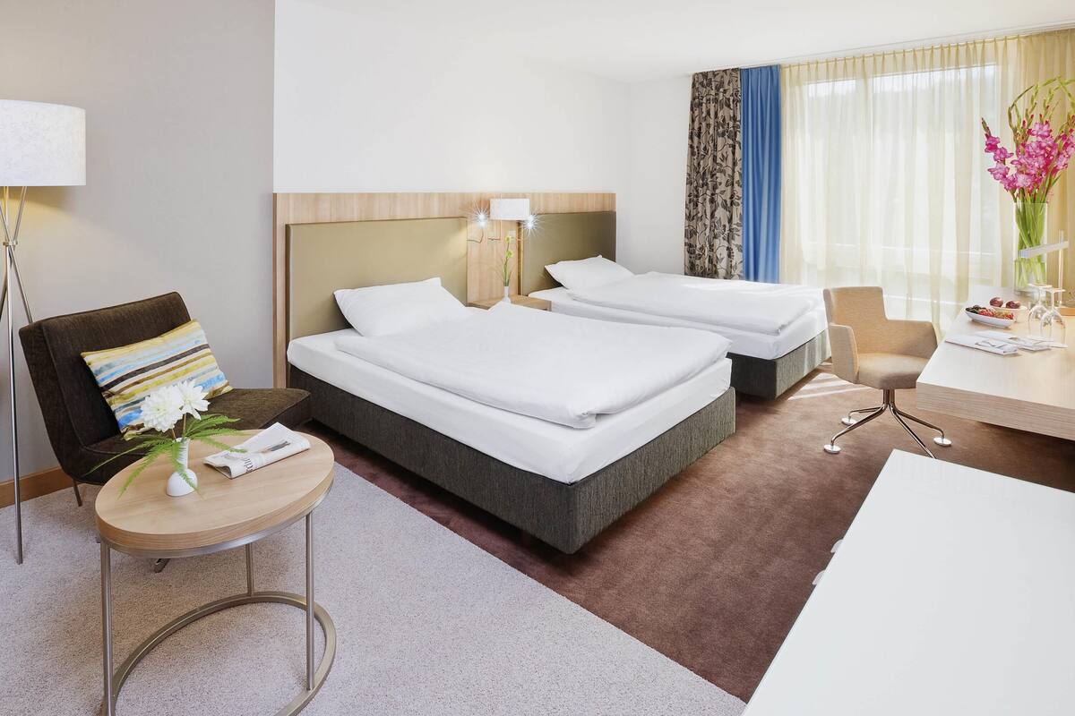 Une chambre d’hôtel avec deux lits, un bureau, une chaise, une lampe et un vase de fleurs.