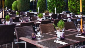 Un espace de restauration en plein air avec des tables disposées pour deux personnes, chacune agrémentée d’une plante en pot, de verres à vin et de couverts.