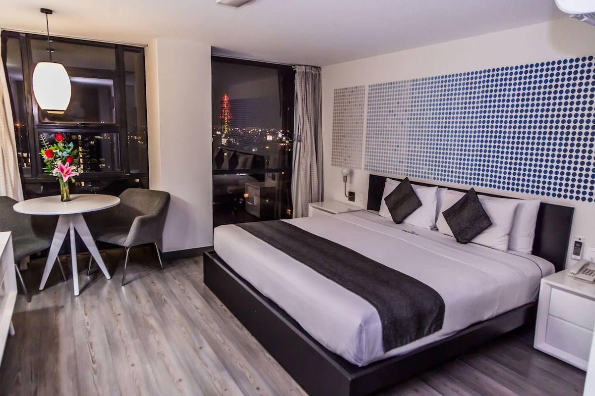 Ein Hotelzimmer mit Bett, Nachttisch, kleiner rundem Tisch mit Stühlen, einer Vase mit Blumen und Blick auf eine nächtliche Stadtlandschaft.